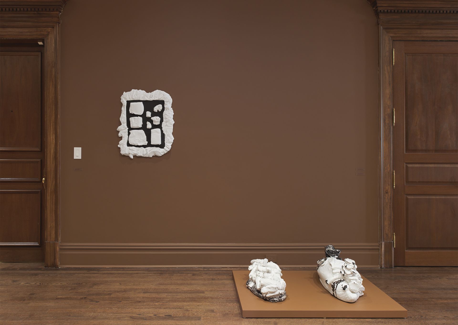 Two white sculptural objects on a brown platform with a white wall-mounted artwork featuring a black wooden plate with white shapes assembled on it.