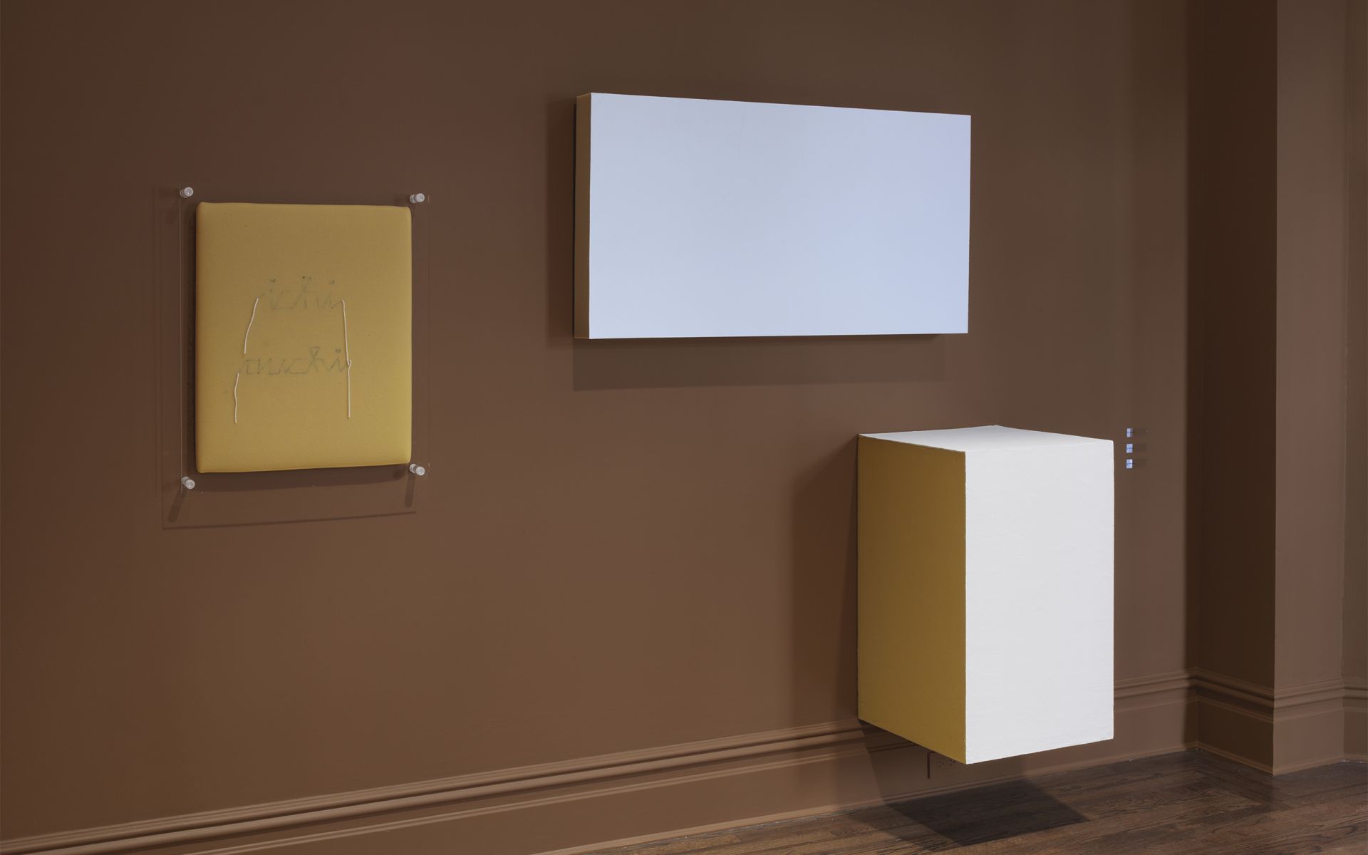 Gallery wall with brown background featuring multiple artworks: yellow work in clear frame, white rectangular work installed horizontally, and white/yellow geometric cube structure mounted at different heights.