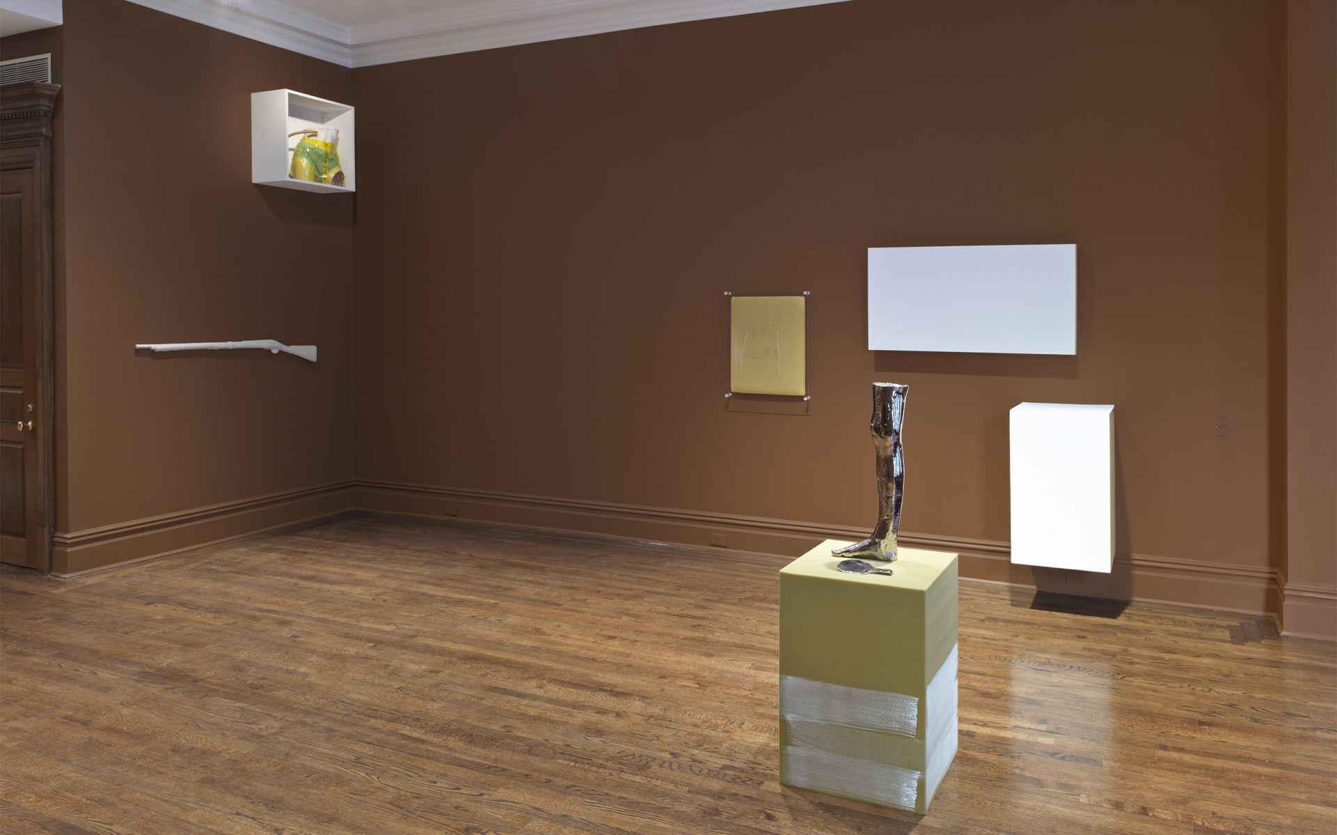 Contemporary gallery space with brown walls displaying white rifle sculpture, mounted rectangular artworks, and metallic sculpture resembling a leg on yellow pedestal. Wooden flooring and molding complete the exhibition space.