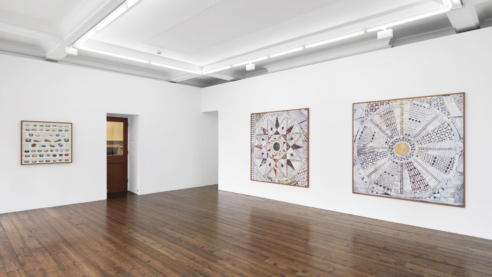 Gallery view showing two large circular mosaic artworks and ceramic collection displayed on white walls with wooden floors
