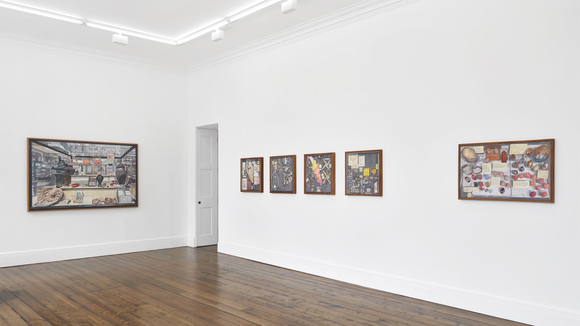 Gallery installation view showing framed artworks on white walls including paintings and a ceramic collection display