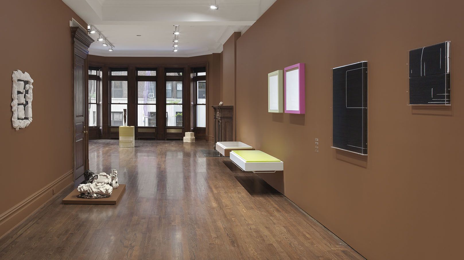 Gallery space with brown walls, track lighting, wood flooring, and bay windows. Features wall-mounted artworks including green and pink frames, black pieces, and two floating platform beds.