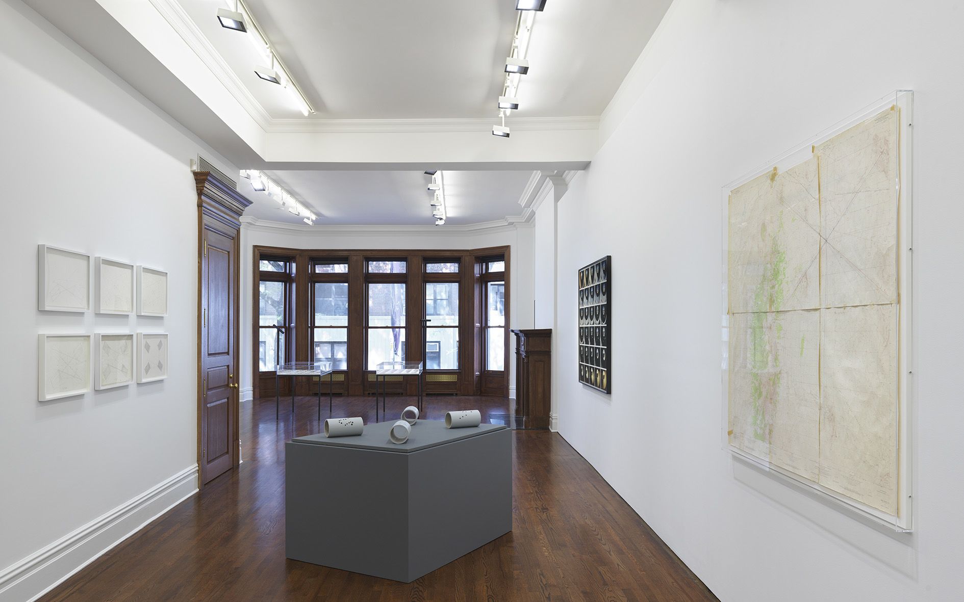 Contemporary art gallery with white walls, dark wood floors, and a bay window. Features white ceramic sculptures on a gray pedestal and framed works.
