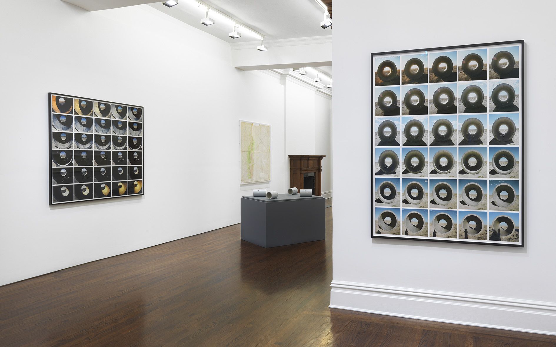 Gallery space displaying two large grid photographic artworks on white walls, with ceramic sculptures on a central gray platform.