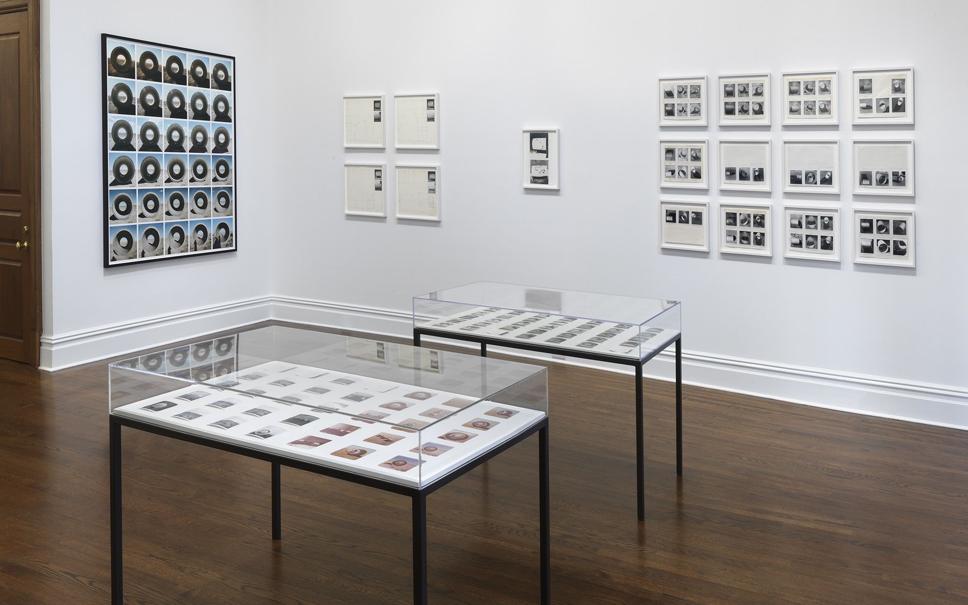 Gallery corner showing photographic grid work alongside smaller framed pieces and glass display cases containing archival materials.