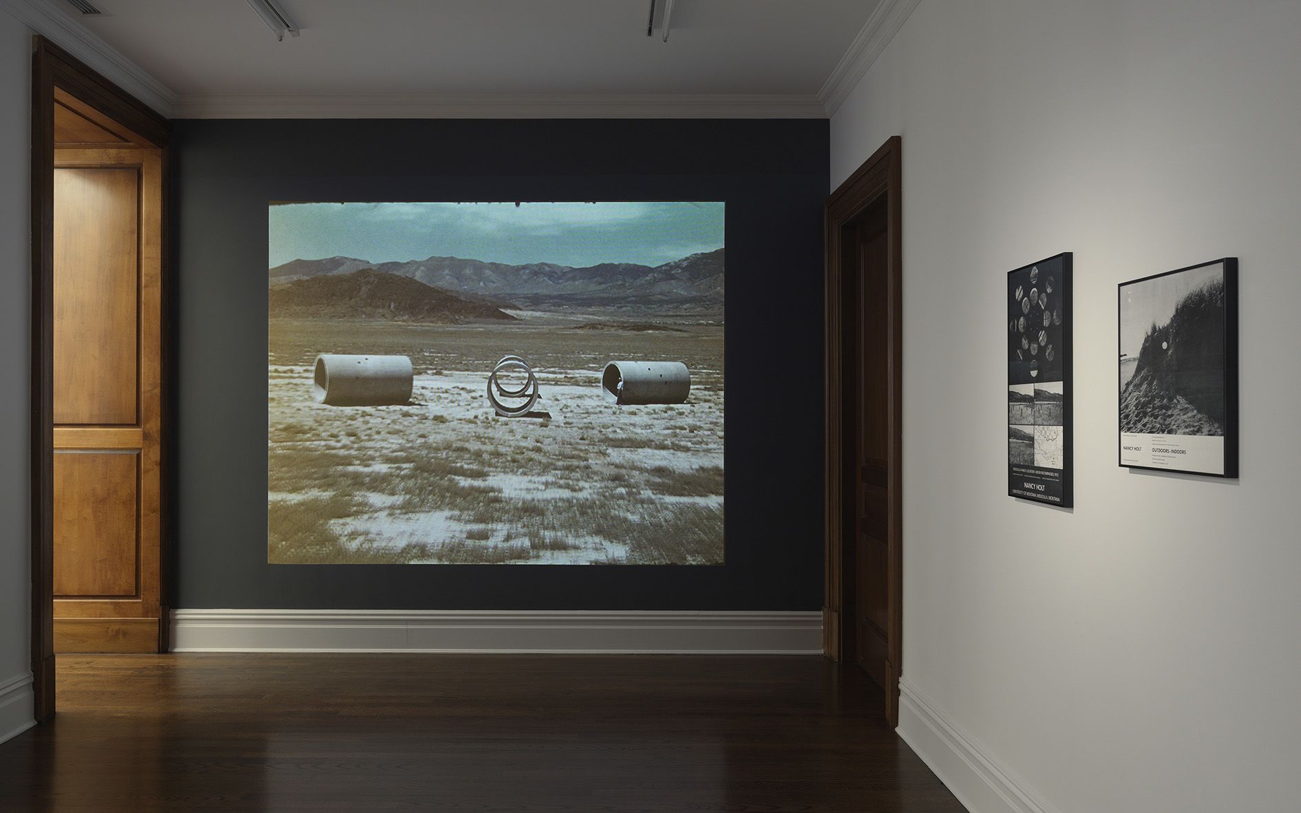 Gallery room with dark accent wall displaying a large video projection, with smaller framed works on adjacent white walls.