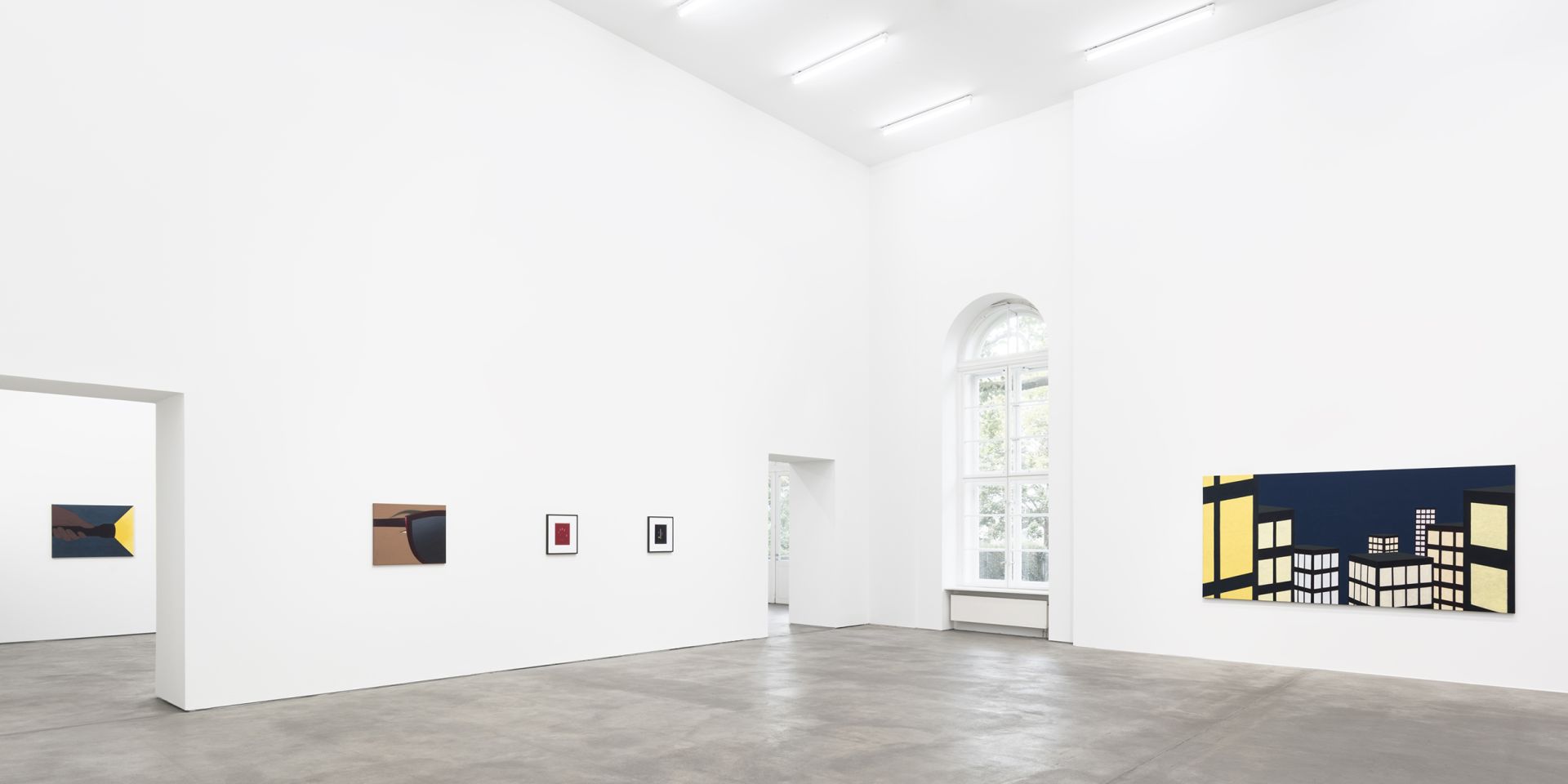 White gallery space with various artworks on walls including geometric paintings and a large nighttime cityscape with illuminated buildings.