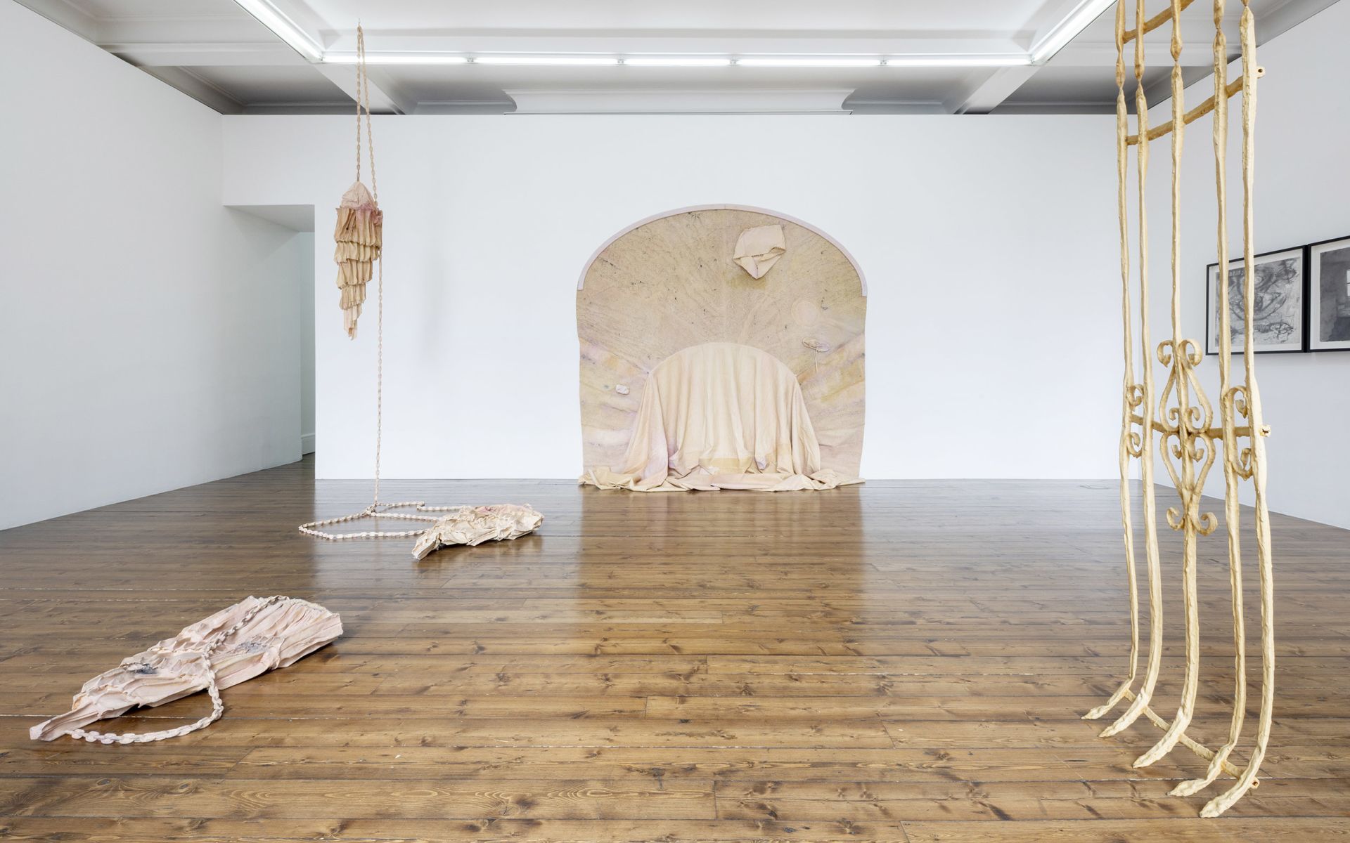 Gallery space with arched alcove containing draped fabric sculpture, hanging textile pieces, and latex barrier installation.
