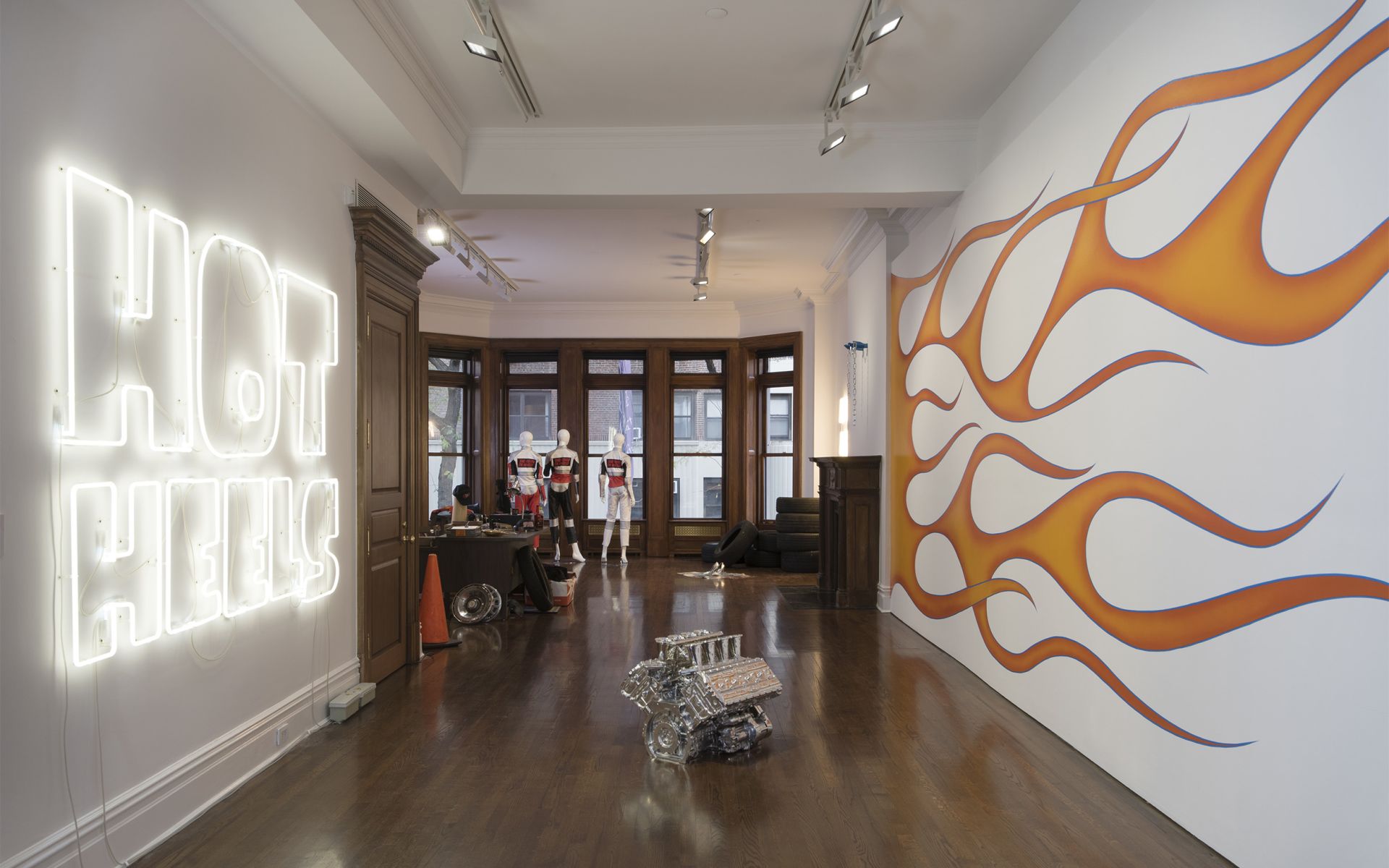 Gallery space with neon "HOT HILLS" sign, orange flame mural, engine on floor, and racing suits on display by windows.