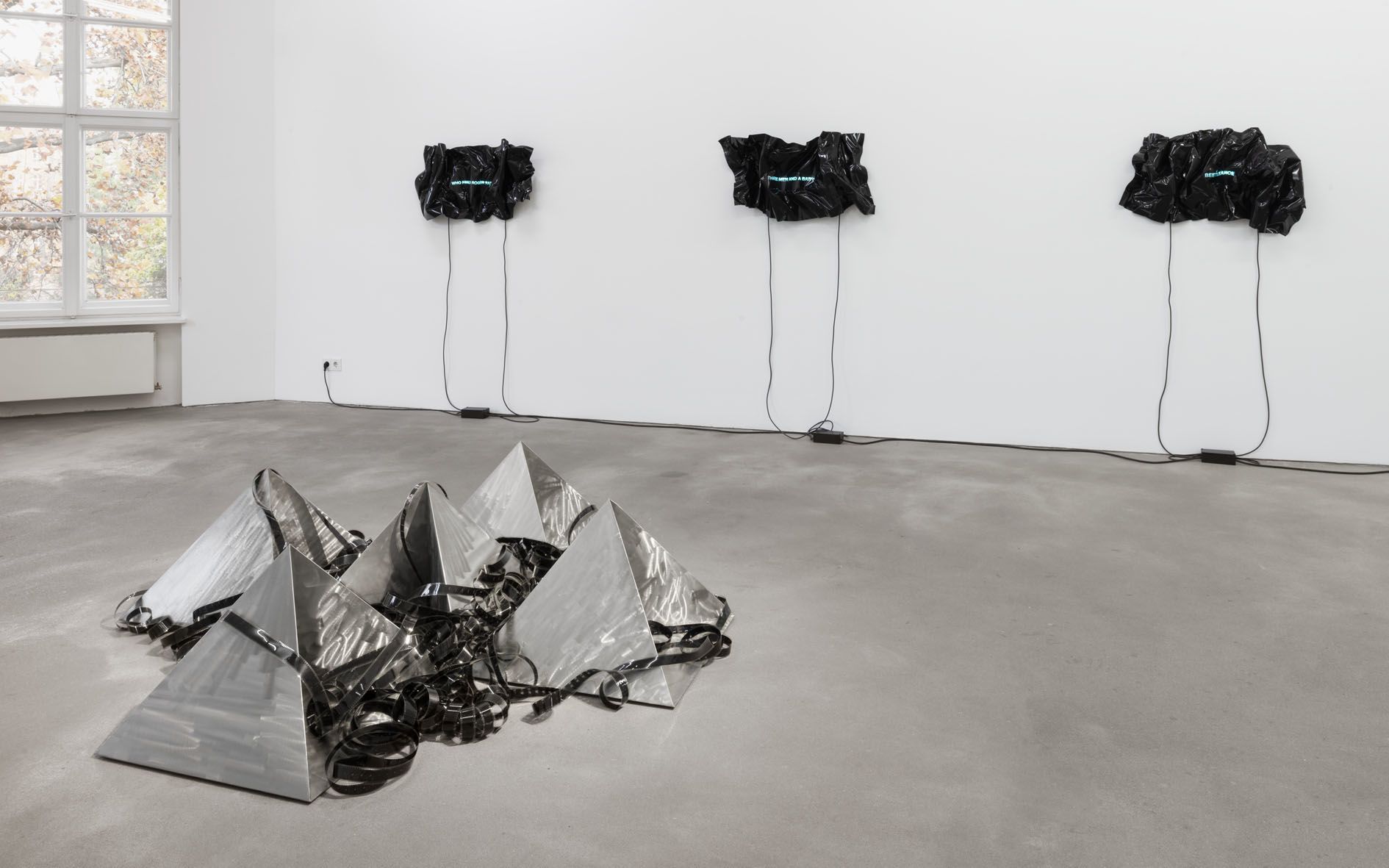 Gallery space with three wall-mounted black vinyl sheets with neon text and reflective pyramids on floor.