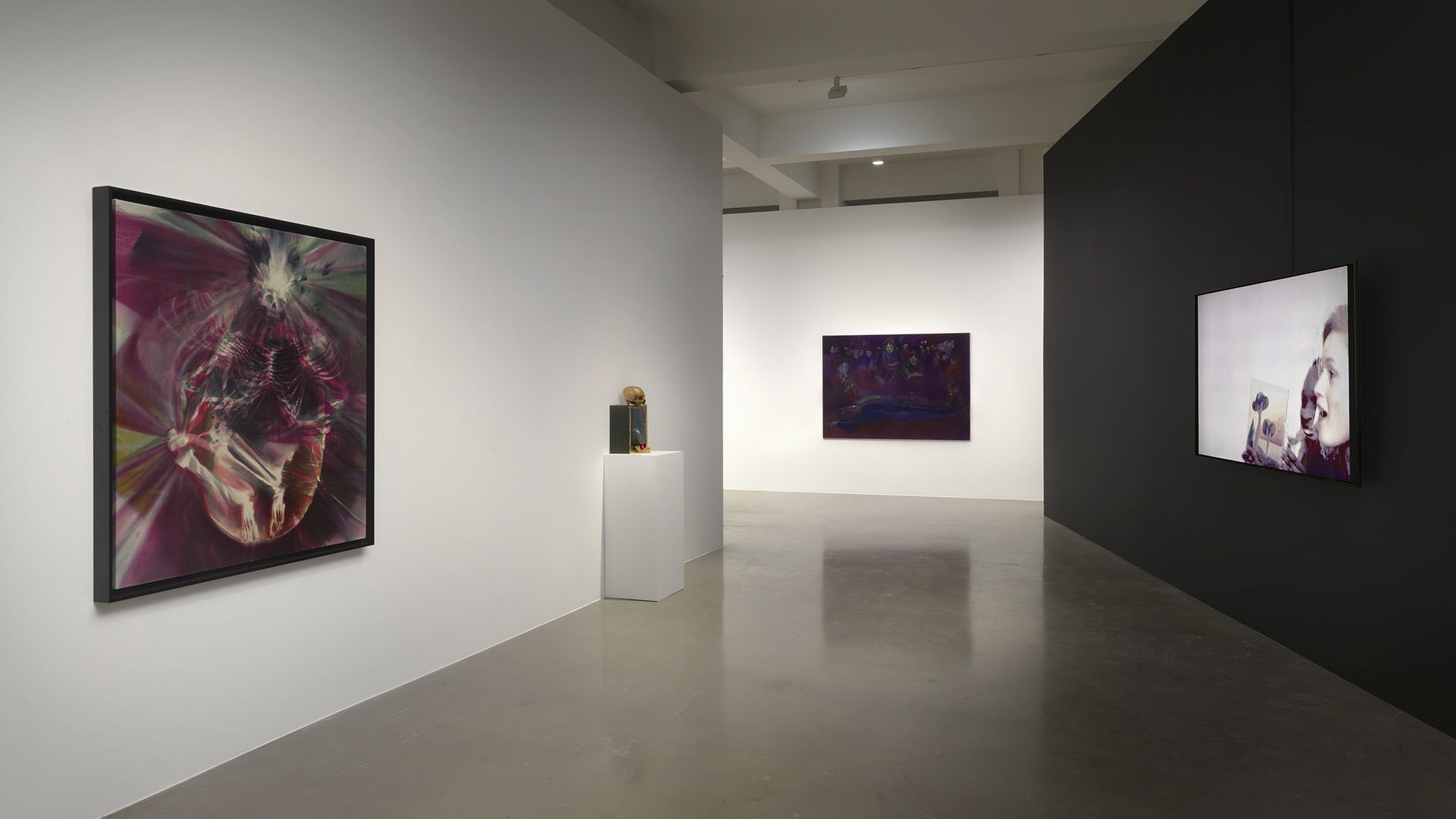 Contemporary art gallery space displaying framed digital and video artworks on white and black walls, with a sculptural object on a pedestal.