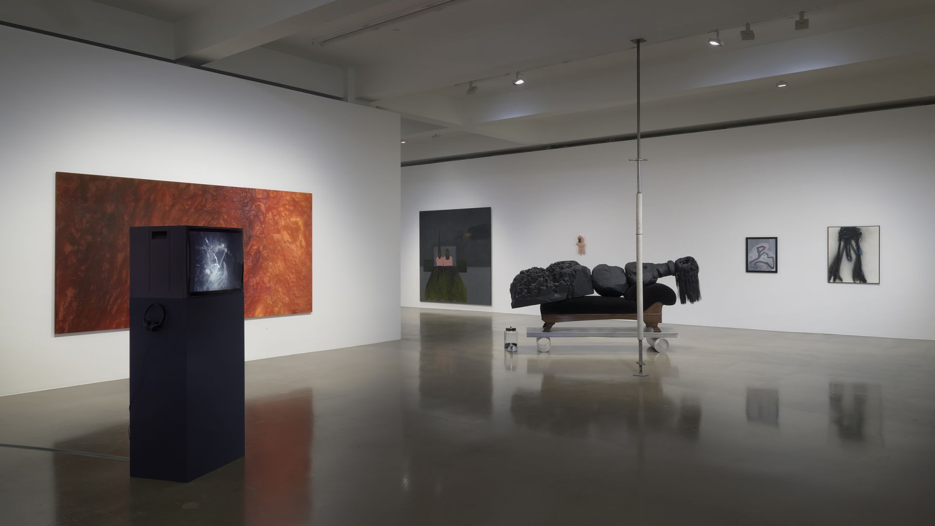 Contemporary art gallery interior with large rust-red abstract painting, black sculptural pedestal, reclining figure with black hair on platform, and smaller framed artworks on white walls.
