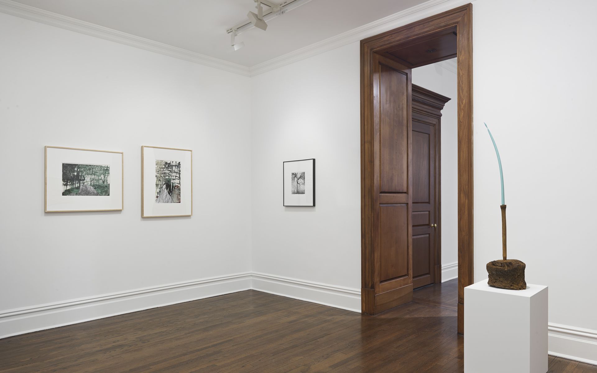 Gallery corner with three framed works and tall sculptural object on pedestal by doorway
