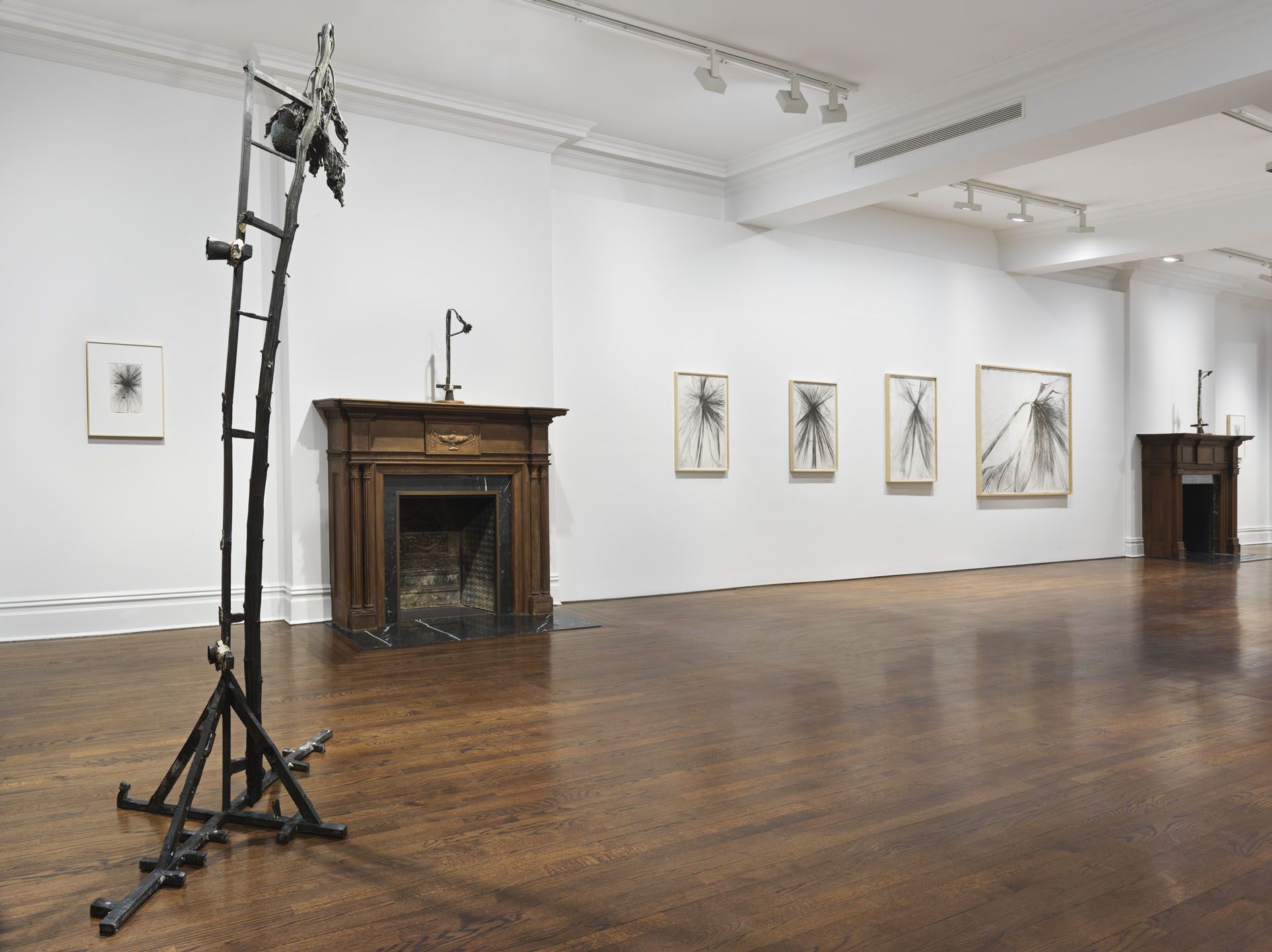 Gallery view with ladder sculpture, framed graphite drawings on wall, and wooden fireplace