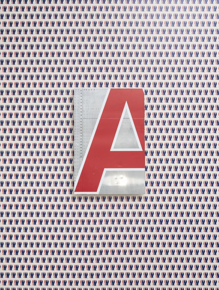 Close-up of a wall covered in repeating blue and red pattern wallpaper with a large red letter "A" mounted on an aluminum backing