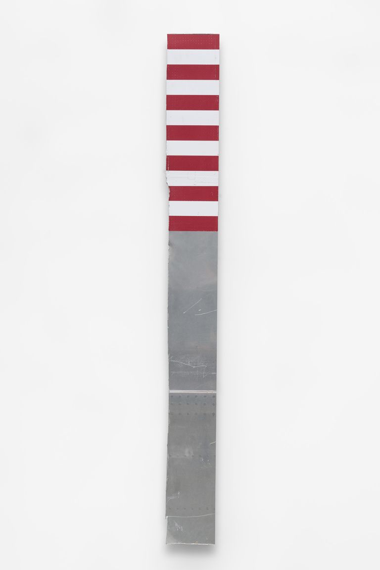 Vertical sculpture or art piece with red and white stripes on the upper portion and gray metallic surface on the lower half, mounted against a white wall