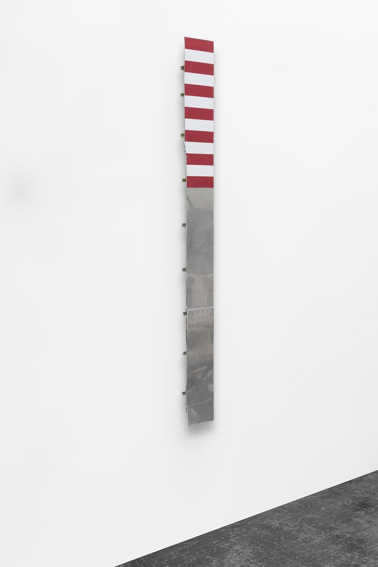 Vertical sculpture or art piece with red and white stripes on the upper portion and gray metallic surface on the lower half, mounted against a white wall