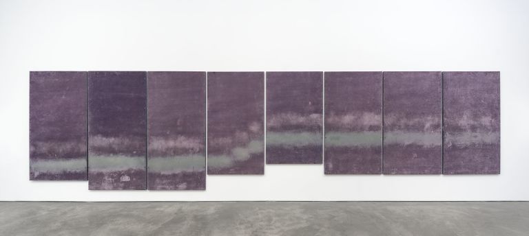 Eight purple abstract paintings arranged in a horizontal line on a white gallery wall, featuring cloudy, atmospheric textures.