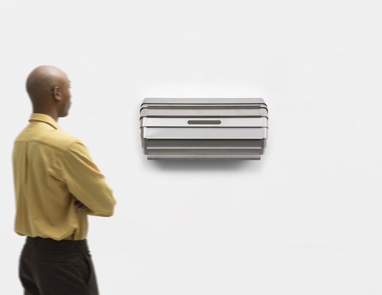 Geometric metal wall sculpture with layered angular forms in silver/gray finish, creating dimensional shadow patterns.