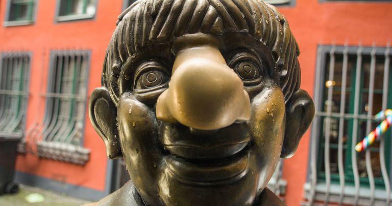 Close-up of bronze or brass sculpture head with exaggerated facial features, large nose, and stylized hair against orange building background.