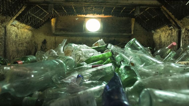 Large pile of empty glass bottles accumulated inside a concrete container or dumpster, illuminated by overhead lighting.