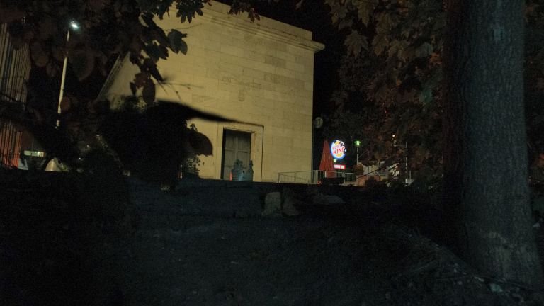 Nighttime view through trees showing an illuminated building facade with a doorway visible in the background.