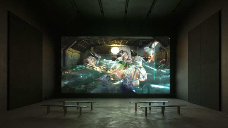 Dark screening room with large projection showing underwater scene of people in diving equipment, wooden benches for seating.