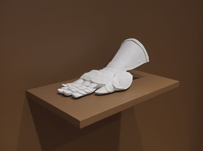 A white sculptural object resembling a partial foot or glove with defined toe or finger sections, displayed on a light brown pedestal against a brown background.