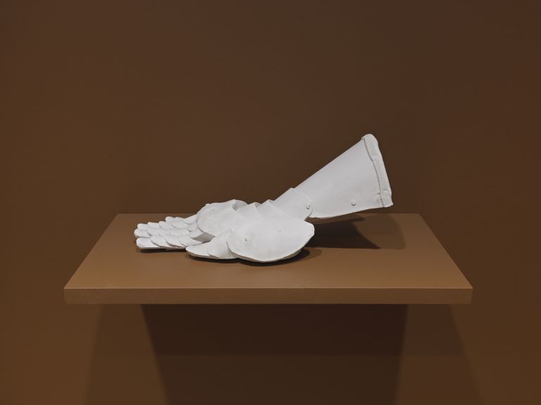 A white sculptural object resembling a partial foot or glove with defined toe or finger sections, displayed on a light brown pedestal against a brown background.