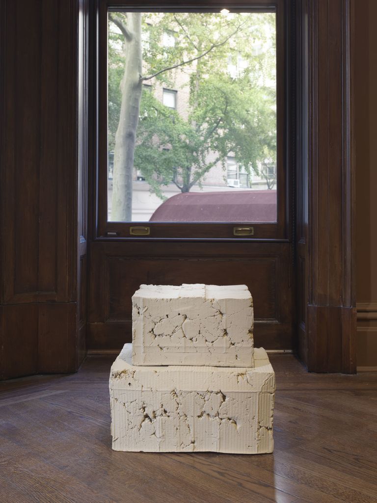 View through ornate wood-framed window with trees visible outside. A sculptural work consisting of two stacked white ceramic blocks sit on wooden floor in window alcove.