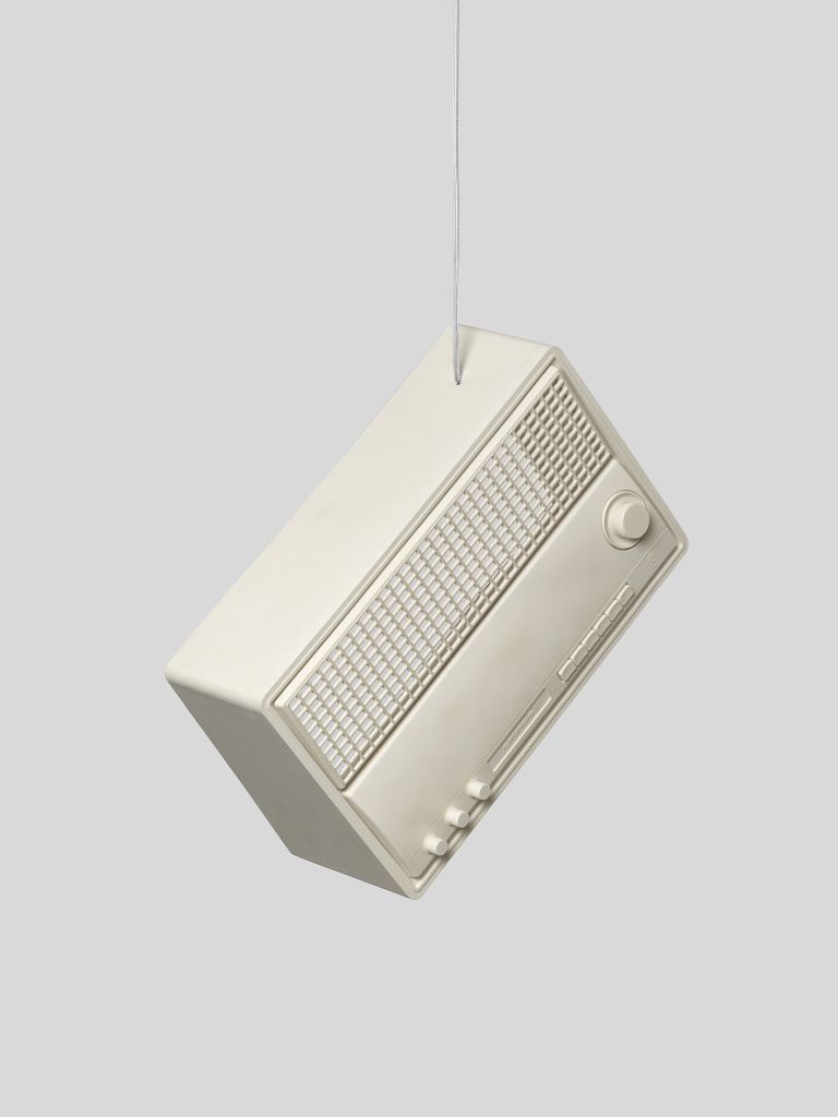 A vintage cream-colored sculpture of a radio suspended by a thin wire against a light gray background. The radio has a rectangular design with a speaker grille, control knob, and several buttons.