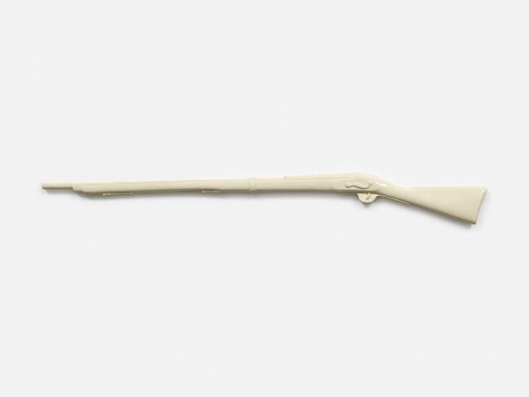 Cream-colored ceramic rifle sculpture mounted on white wall, displayed horizontally as an art piece against minimalist background.