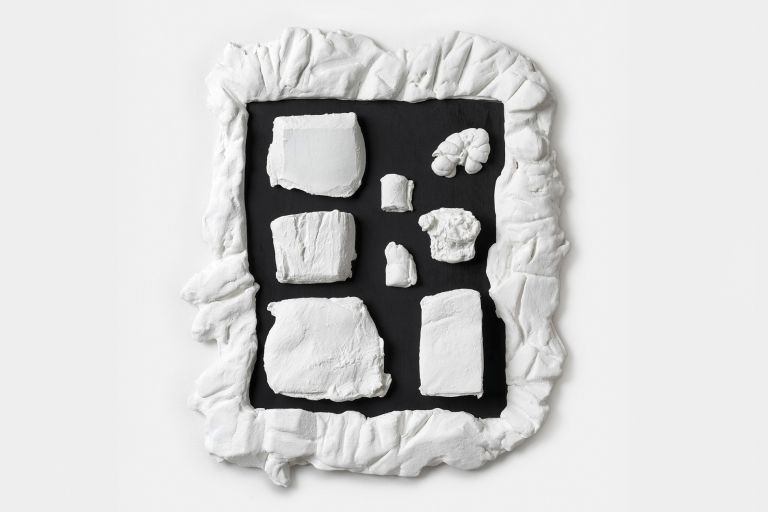 A white-framed black panel with various white sculptural fragments arranged in a grid pattern, resembling an artistic specimen display or collection.