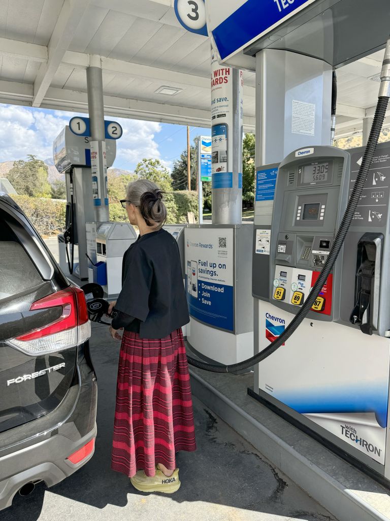 Person in plaid skirt and dark top standing at gas station pump, filling vehicle tank during daytime performance documentation.