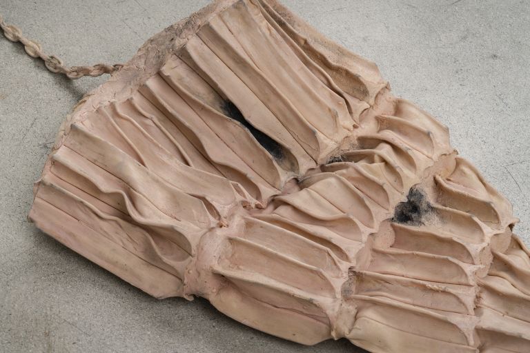 Sculptural object resembling layered fabric or paper with braided rope attachment on concrete floor.