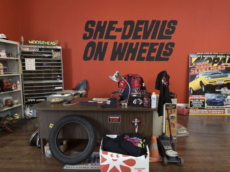 Red wall with "SHE-DEVILS ON WHEELS" text above cluttered desk with tire, racing gear and memorabilia.