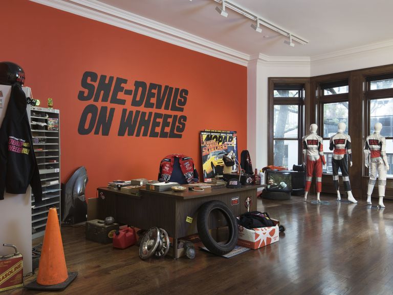 Red wall with "SHE-DEVILS ON WHEELS" text above cluttered desk with tire, racing gear and memorabilia.