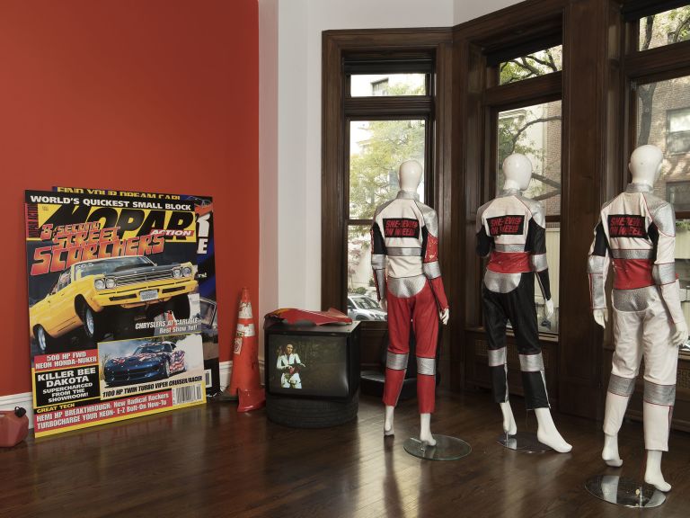 Red wall with "SHE-DEVILS ON WHEELS" text above cluttered desk with tire, racing gear and memorabilia.