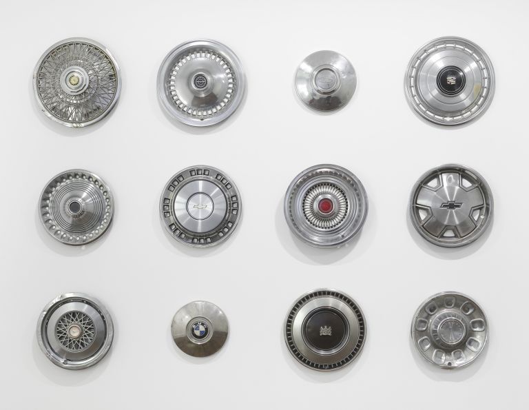 Grid of twelve vintage chrome car hubcaps and wheel covers mounted on white wall.