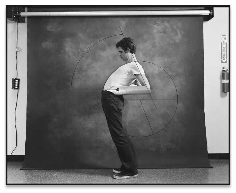 Person in white shirt and dark pants standing against gray backdrop with geometric line overlay resembling golden ratio