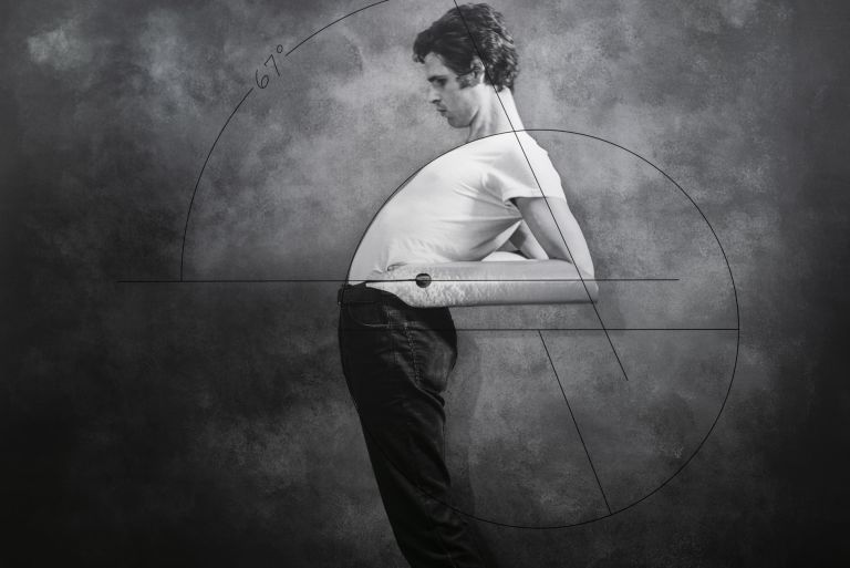 Person in white shirt and dark pants standing against gray backdrop with geometric line overlay resembling golden ratio