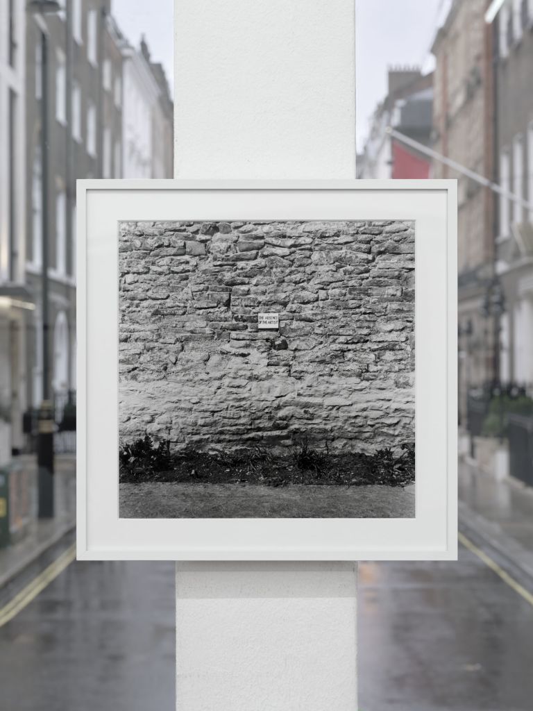 Framed photograph of stone wall with small sign, displayed in window with street view visible in background