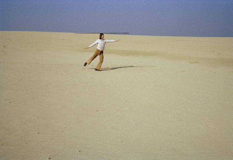 Color photograph of a person in white shirt with arms outstretched walking across a vast sandy desert under blue sky.
