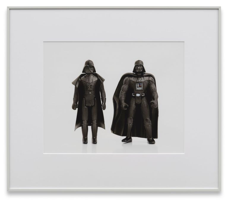 Two Darth Vader action figures of different sizes standing side by side on white background, framed with wide white mat