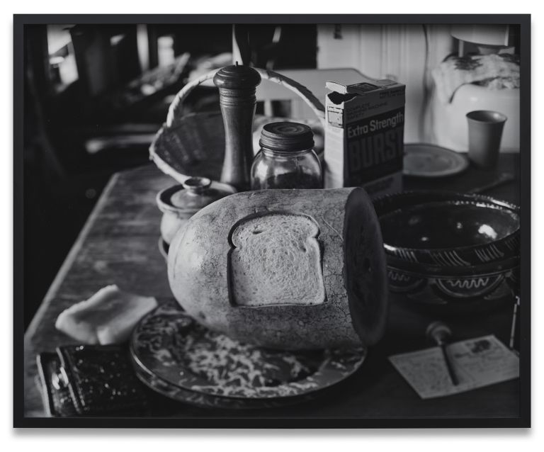 Black and white photo of a watermelon with slice of bread shaped cut-out on its surface, all surrounded by kitchen items including jars, cereal box, and plate.