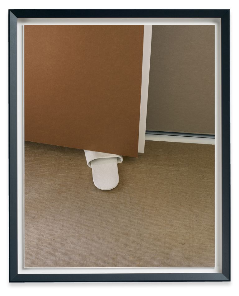 Framed photo showing tongue-like ceramic form protruding from corner where two colored paper sheets meet on wooden surface