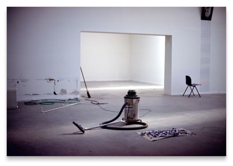 Gallery space with vacuum cleaner, broom, and cleaning supplies on floor beneath white architectural installation