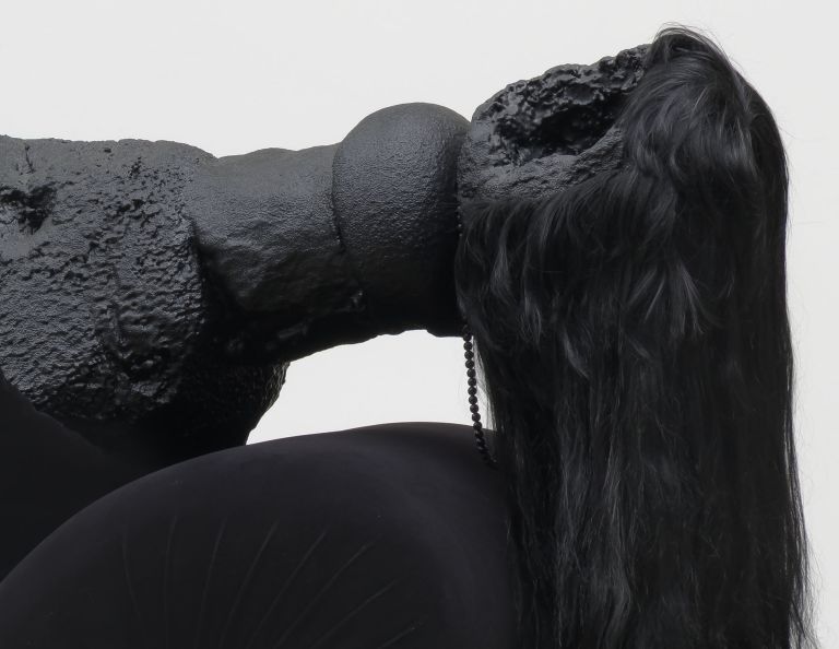 Black sculptural form resembling draped fabric or organic shape mounted on wooden bench and aluminum elements, positioned on gallery floor.