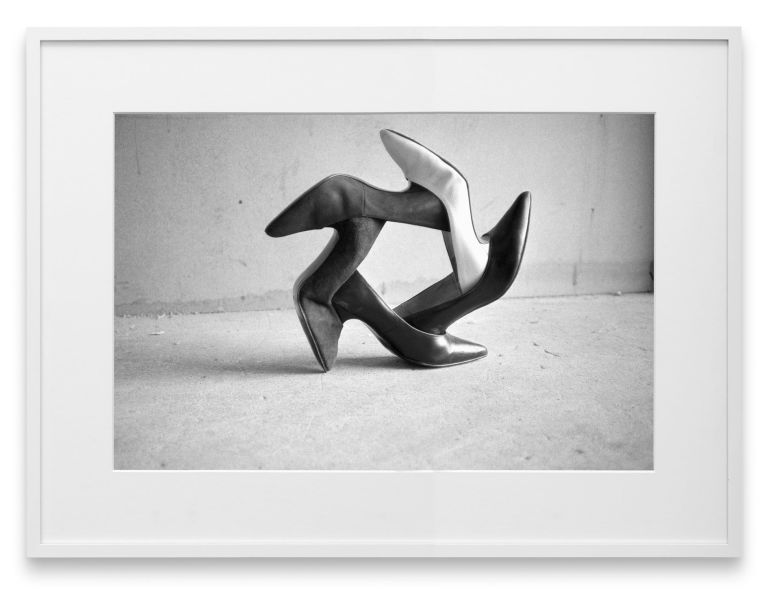Black and white photo of two high-heeled shoes arranged in abstract sculptural formation against plain background