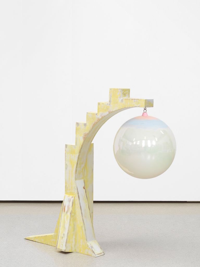 Yellow stepped wooden arch sculpture holding a suspended iridescent white sphere via pink hook.